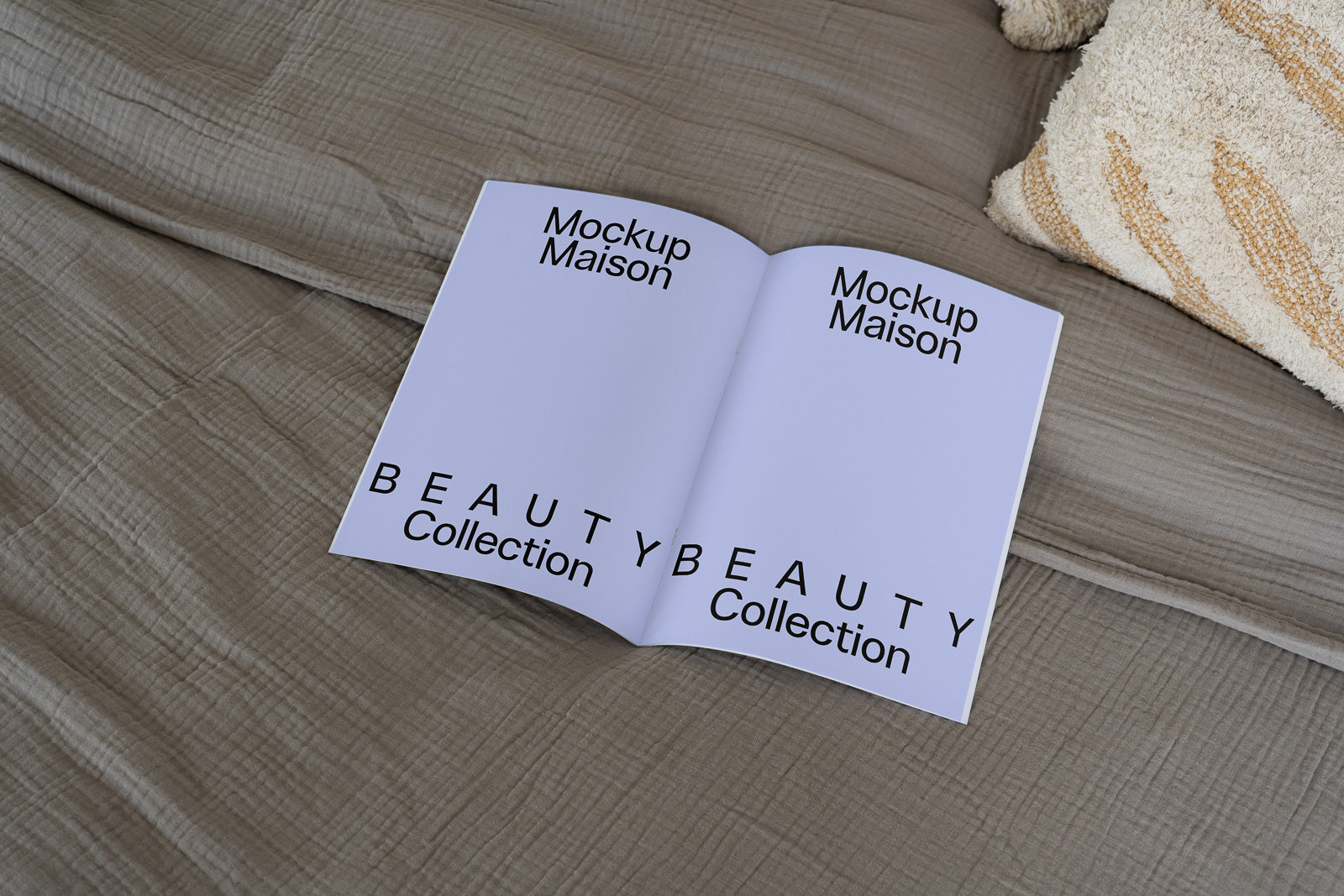 PSD mockup of an open booklet with "Mockup Maison" and "Beauty Collection" text, placed on a beige textured fabric surface near a patterned cushion.
