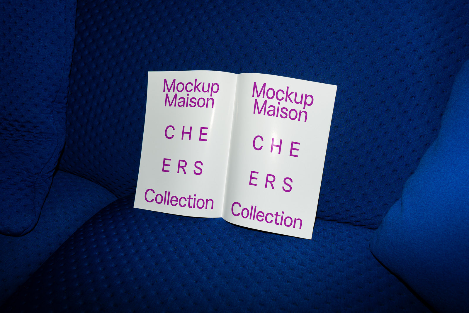 PSD mockup of an open booklet resting on a blue textured surface, displaying the text "Mockup Maison CHEERS Collection" in purple lettering.