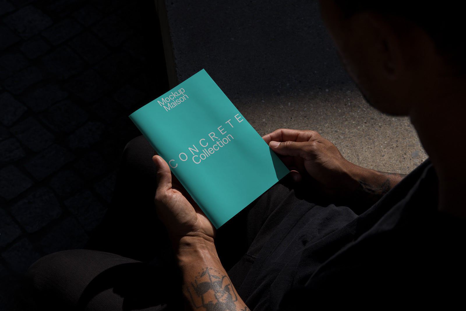 PSD mockup of a person holding a turquoise booklet titled "Concrete Collection" in dim lighting, showing tattoos on their arm.
