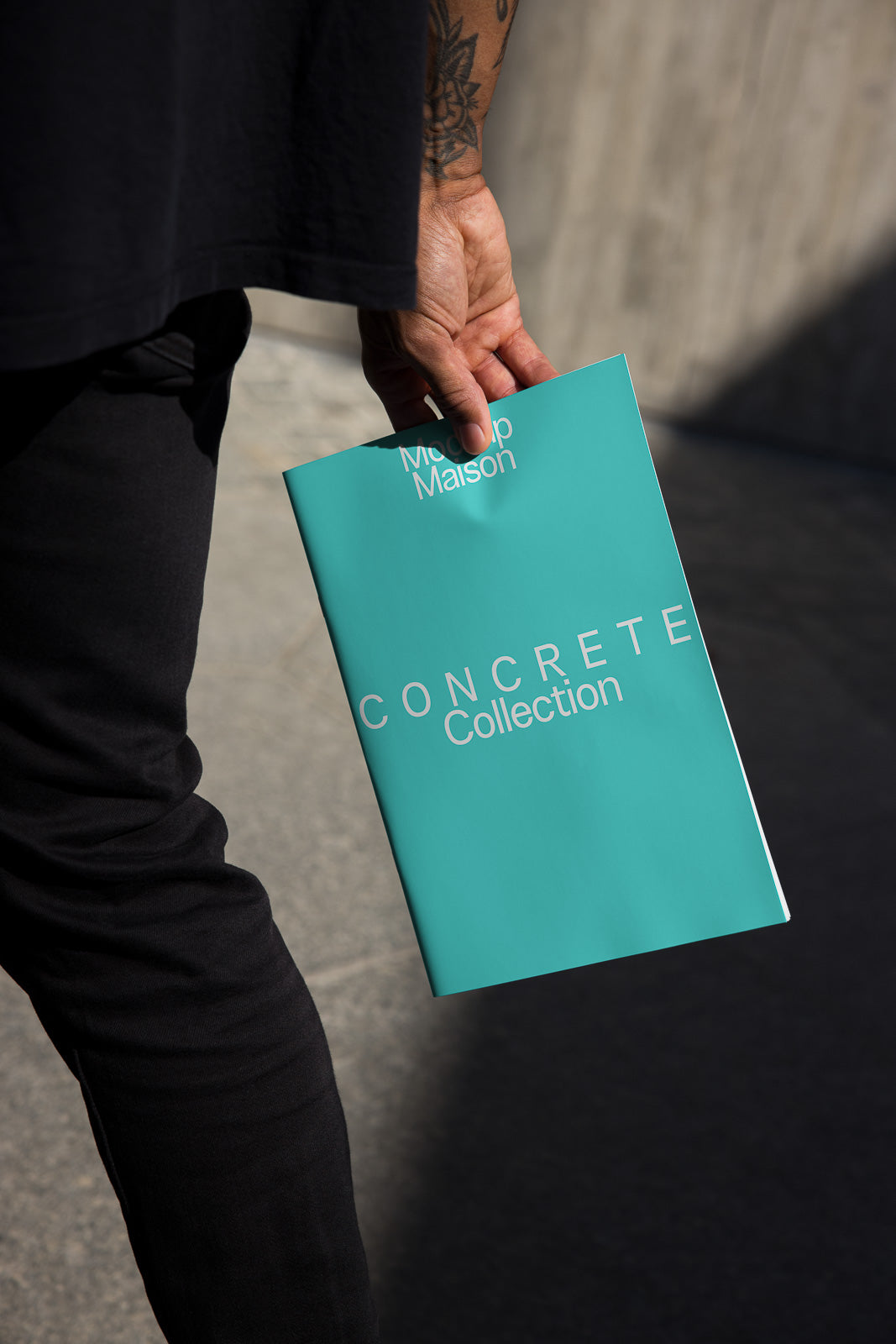 PSD mockup of a person holding a turquoise booklet titled "Concrete Collection" with "Mockup Maison" text, against a blurred urban background.