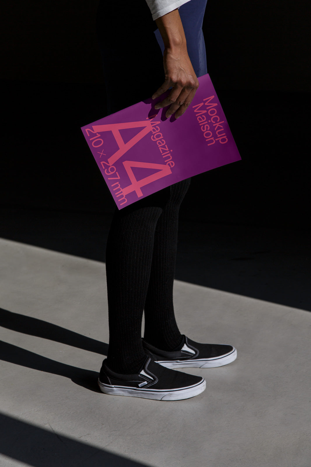 PSD mockup of a person holding a vibrant magenta A4 magazine with a shadow on the floor, wearing black slip-on shoes and long dark socks.