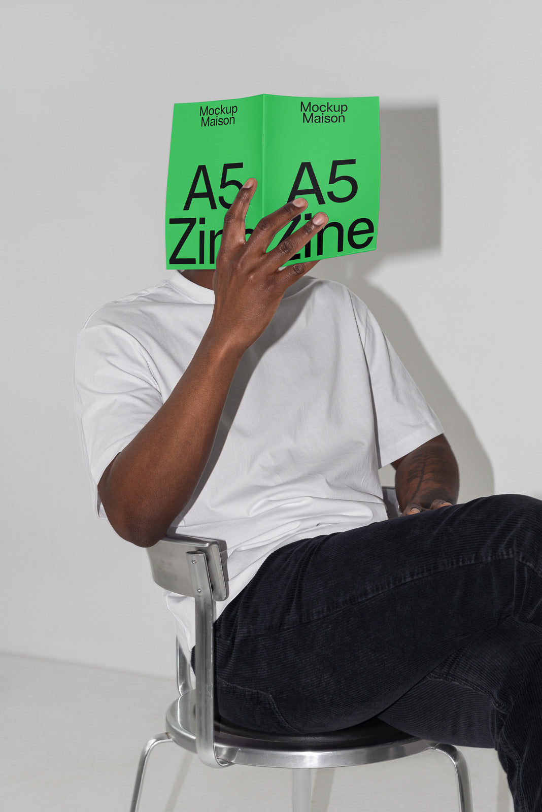 PSD mockup of a person seated in a metal chair, holding a bright green A5 zine covering their face, dressed in a white shirt and dark pants.