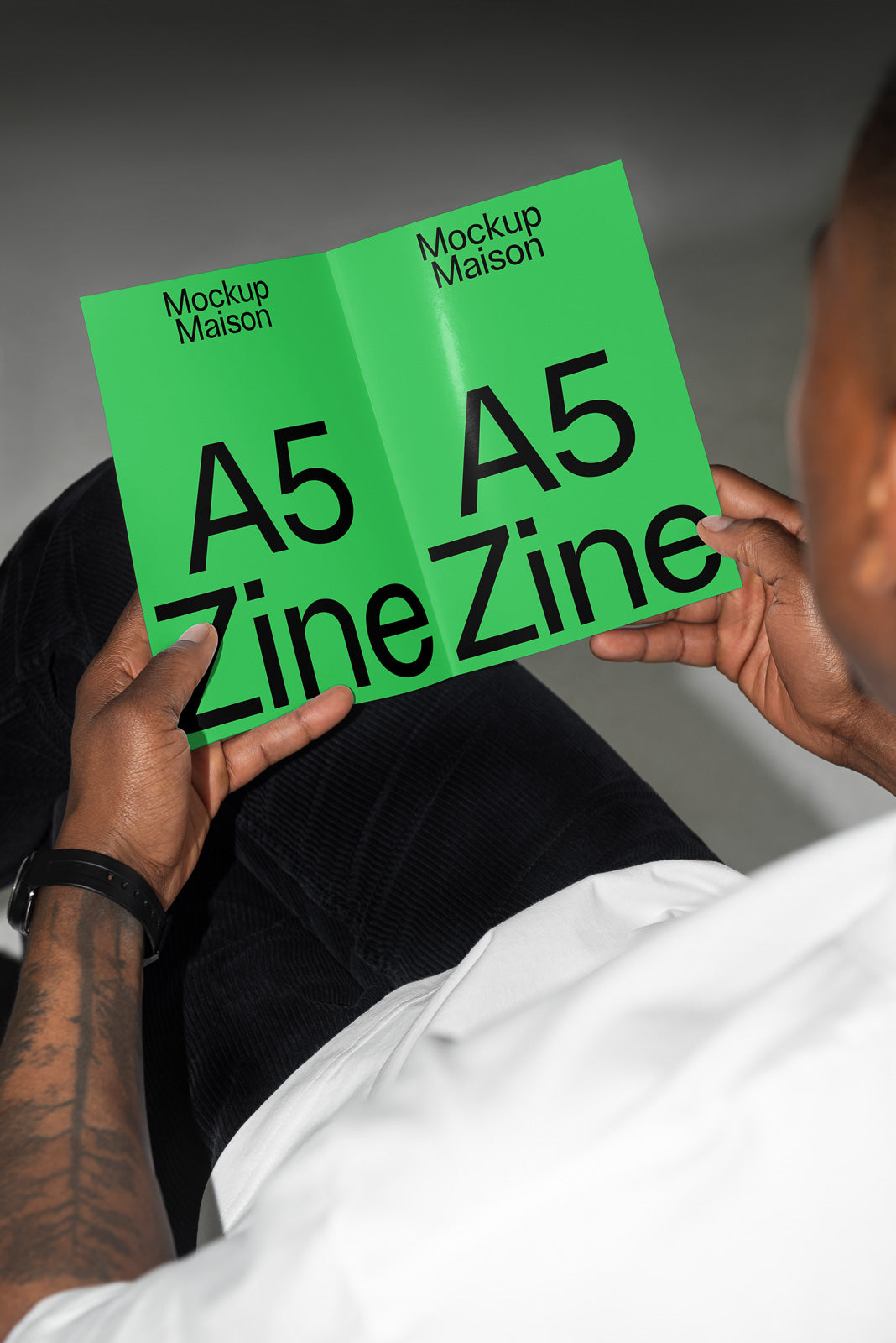 PSD mockup of a green A5 zine with bold black text reading "Mockup Maison A5 Zine," held by a person wearing a white shirt and dark pants.