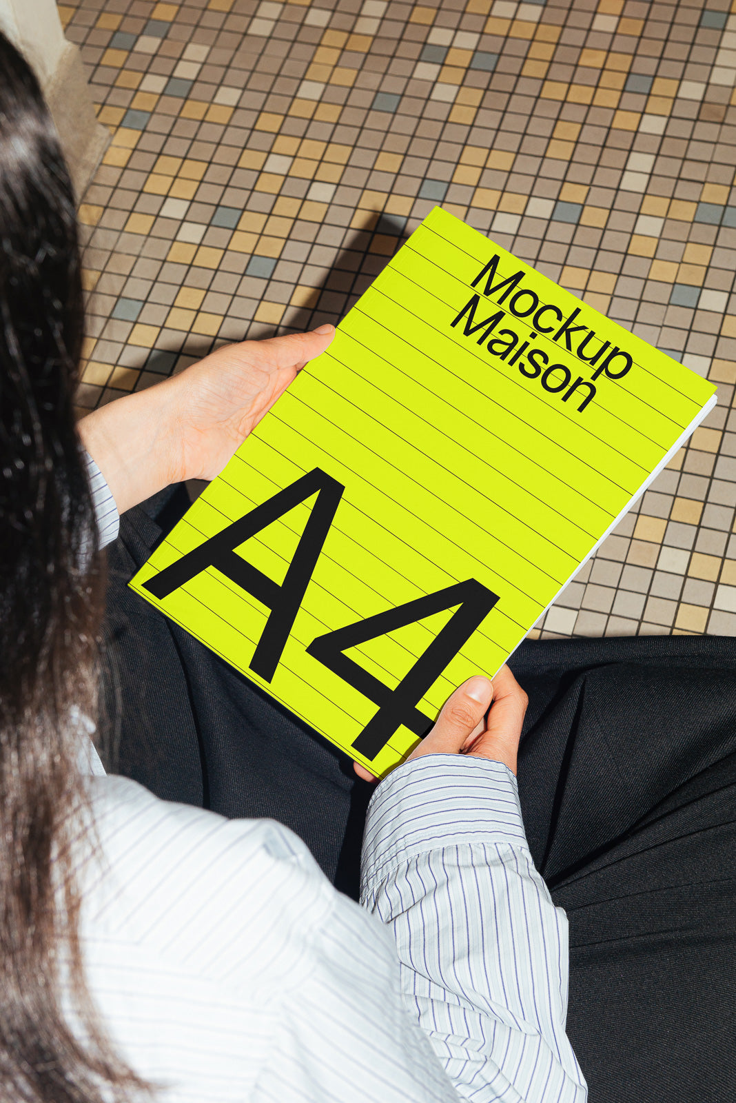 PSD mockup of a person holding an A4-sized bright yellow notebook labeled "Mockup Maison" against a tiled floor background.