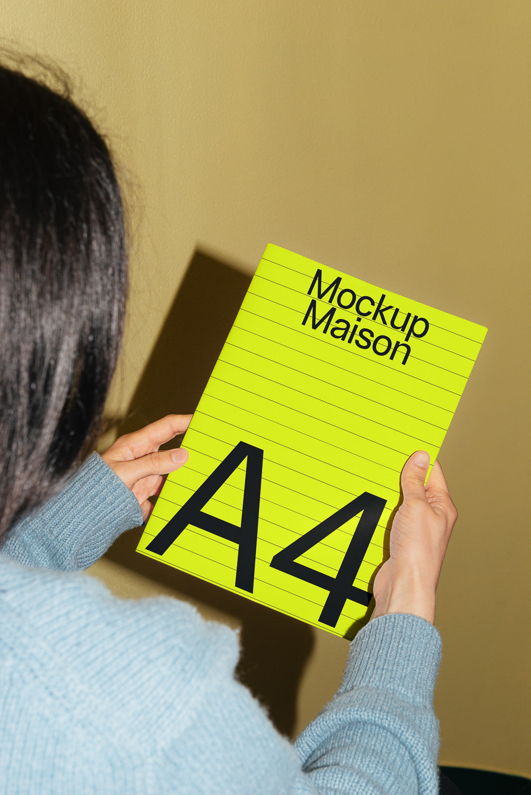 PSD mockup of a person holding a bright yellow A4 sheet titled "Mockup Maison" against a beige background, wearing a light blue sweater.