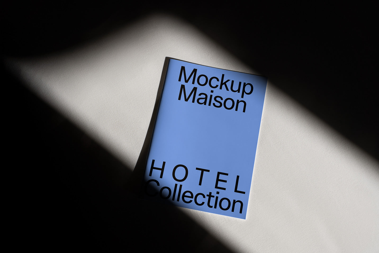 PSD mockup of a blue booklet titled "Mockup Maison HOTEL Collection" illuminated by a focused beam of light on a textured surface.