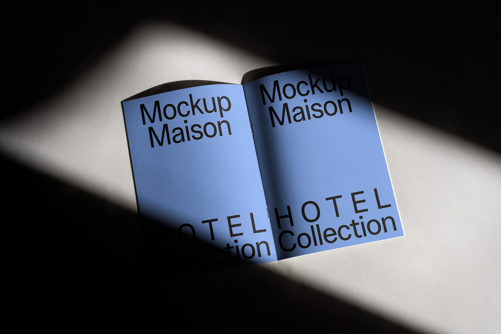 PSD mockup of an open book with a blue cover and bold black text "Mockup Maison HOTEL Collection" illuminated by a beam of light.