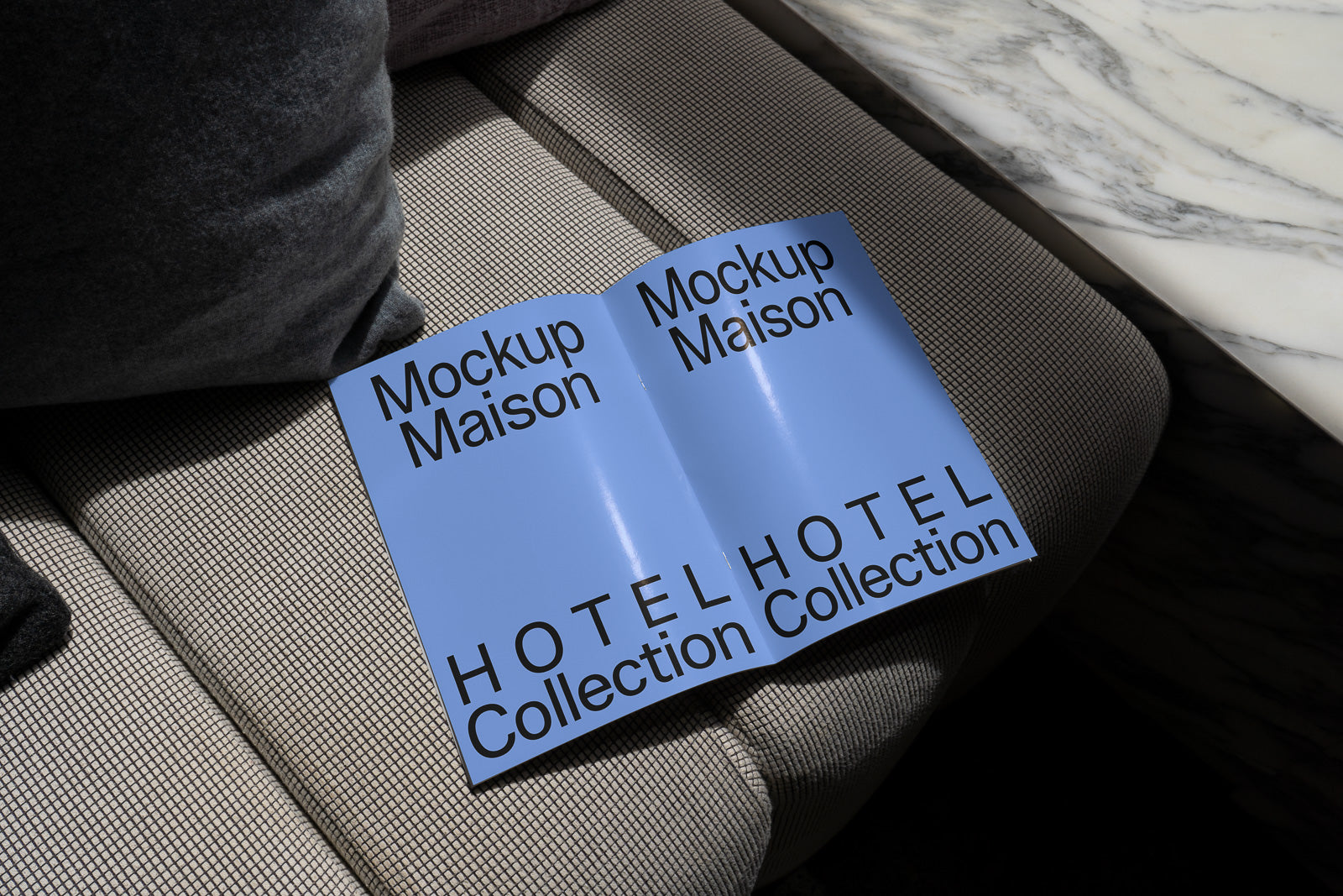 PSD mockup of an open magazine with a blue cover displaying "Mockup Maison" and "HOTEL Collection Collection" on a textured sofa near a marble table.