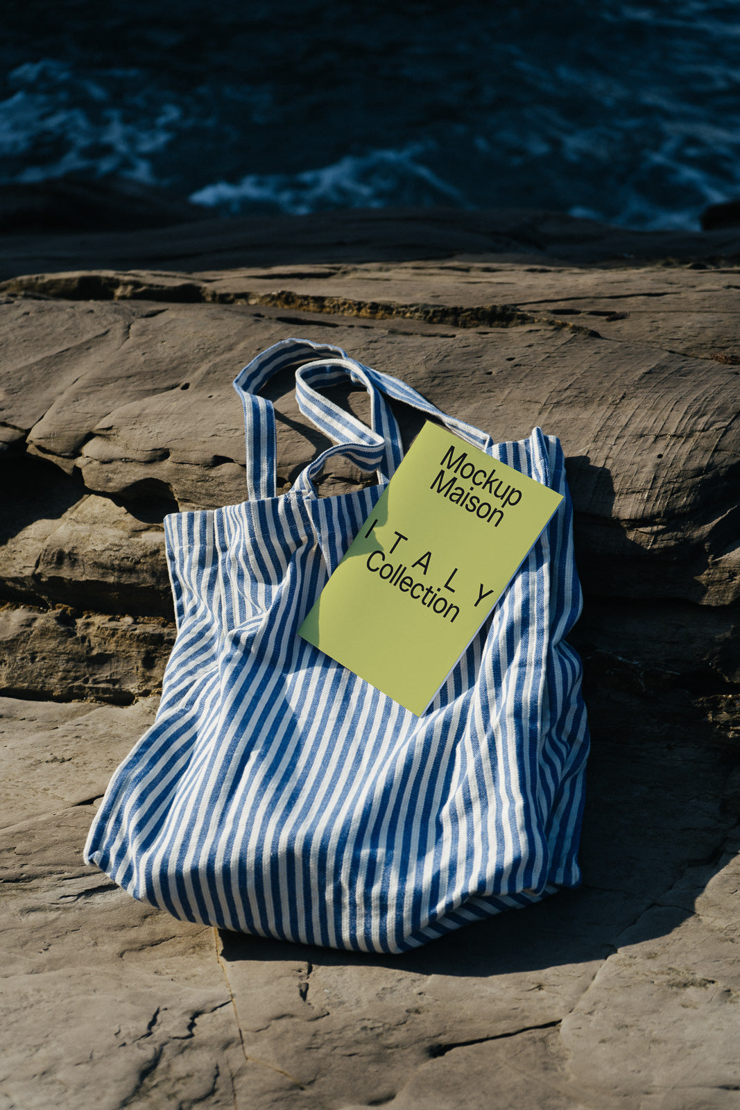 PSD mockup of a striped tote bag with a green card that reads "Mockup Maison ITALY Collection" placed on a rocky surface by the sea.