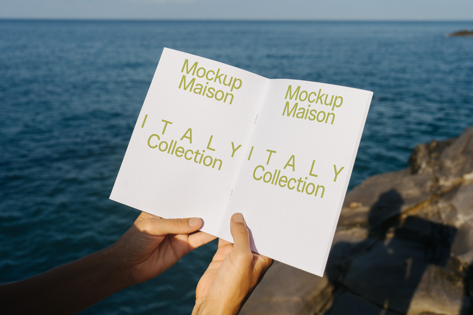 PSD mockup of an open booklet titled "Mockup Maison Italy Collection" held by hands near a rocky shoreline with the sea in the background.