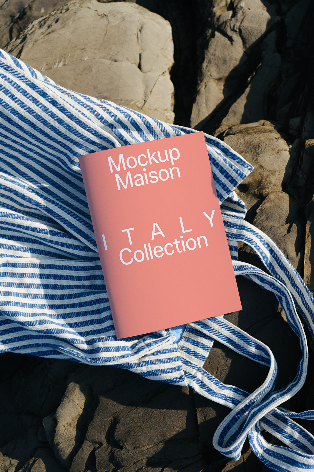 PSD mockup of a pink booklet labeled "Mockup Maison Italy Collection" resting on a blue and white striped fabric over rugged stone.