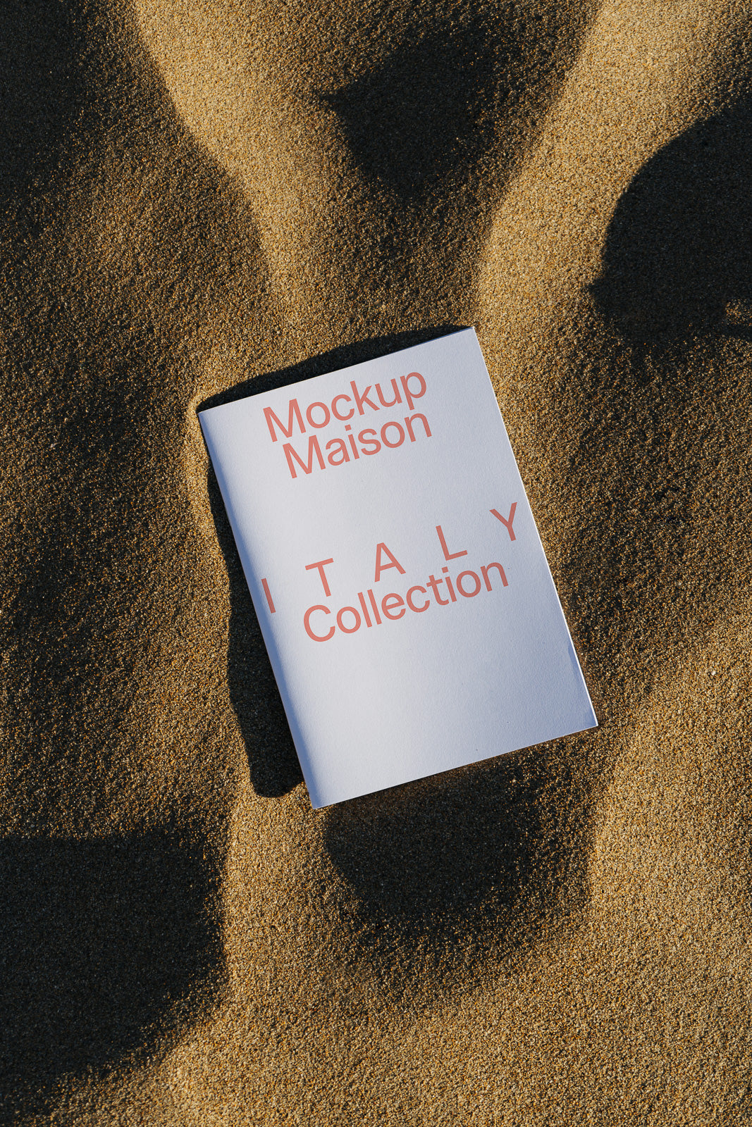 PSD mockup of a "Mockup Maison Italy Collection" booklet on textured sand, casting a shadow with bold red text.