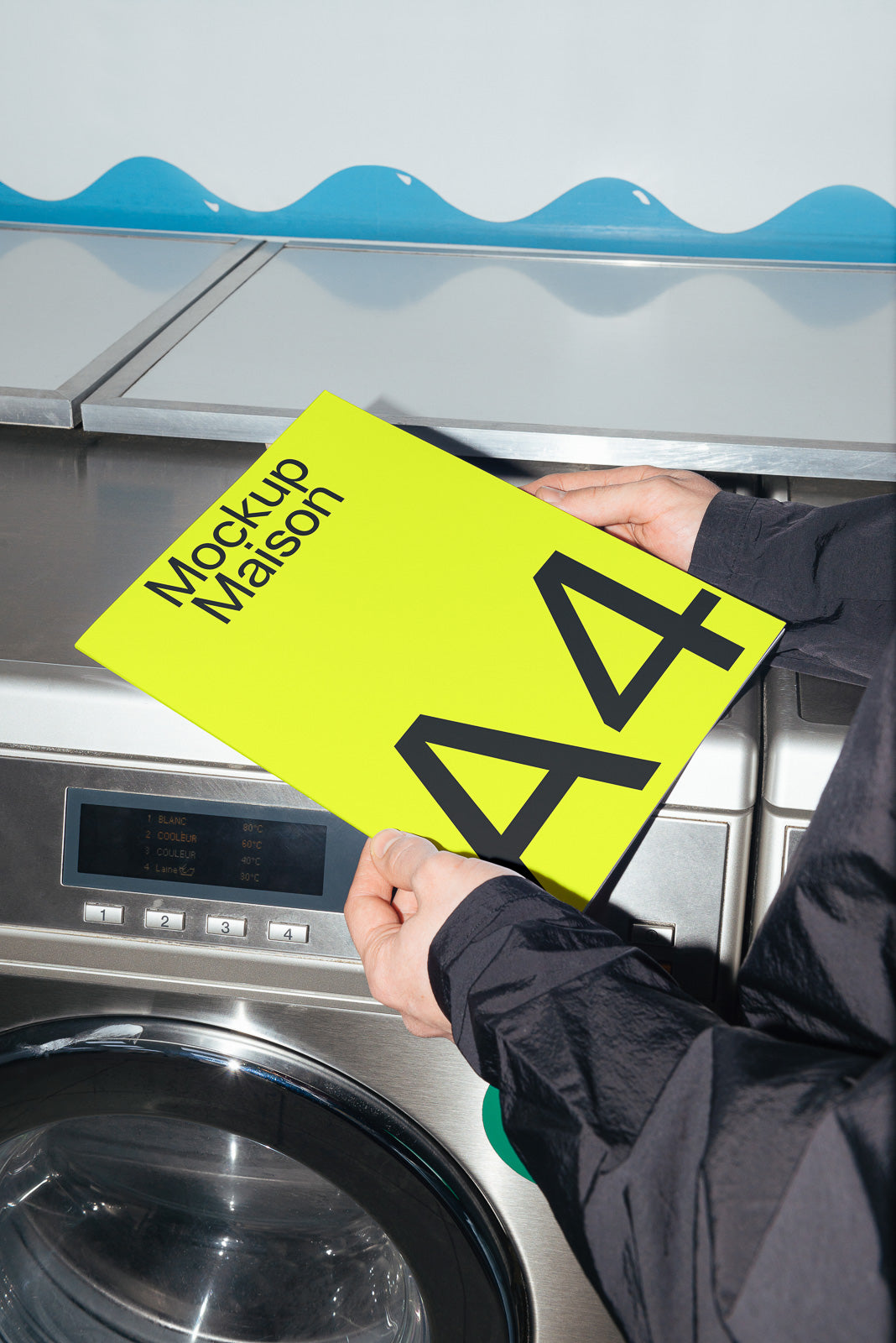 PSD mockup of a vibrant yellow A4 paper with bold black text held by hands above a metallic washing machine, creating an artistic contrast.