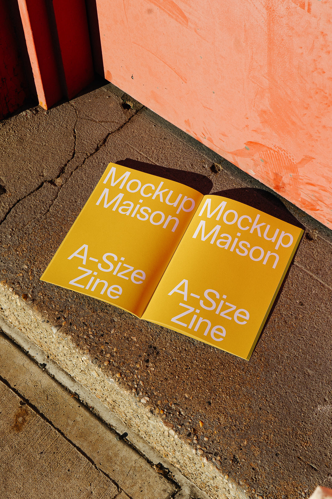 PSD mockup of an open A-size zine with yellow covers, placed on a textured outdoor surface beside an orange wall.