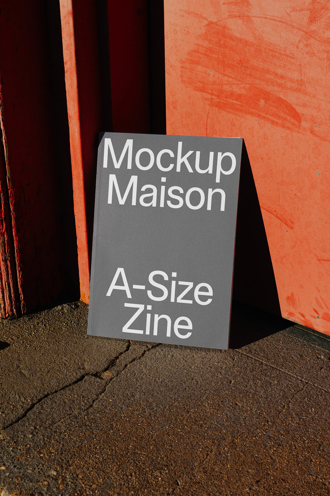 PSD mockup of a gray A-size zine with "Mockup Maison" text, leaning against a red textured wall, casting shadows on the ground.