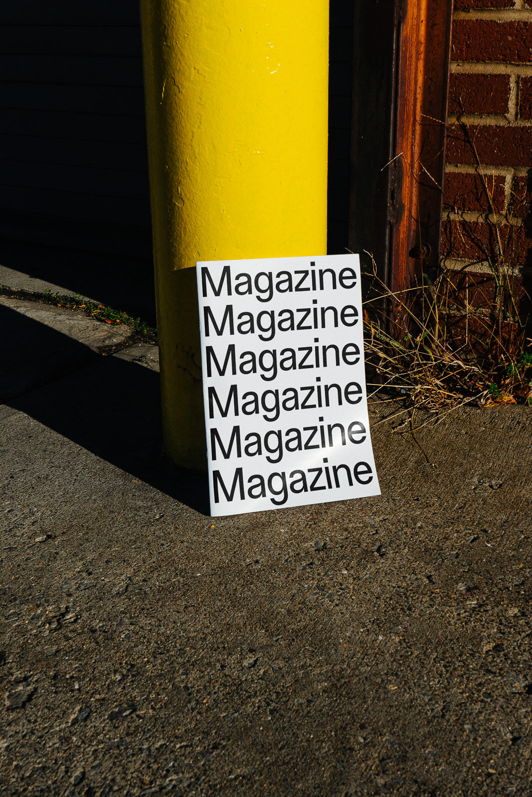 PSD mockup of a magazine cover leaning against a yellow pole, set on a concrete surface with a brick wall and grass in the background.