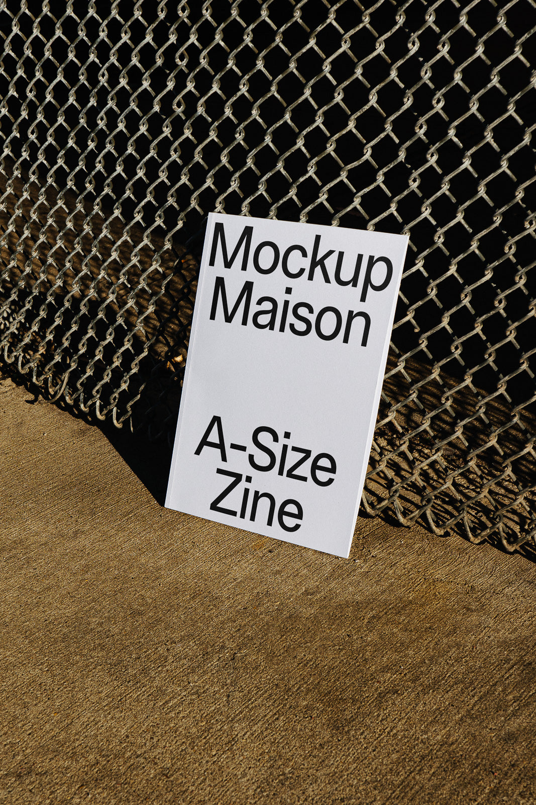 PSD mockup of a vertical A-size zine with "Mockup Maison" text, leaning against a chain-link fence on a concrete surface.