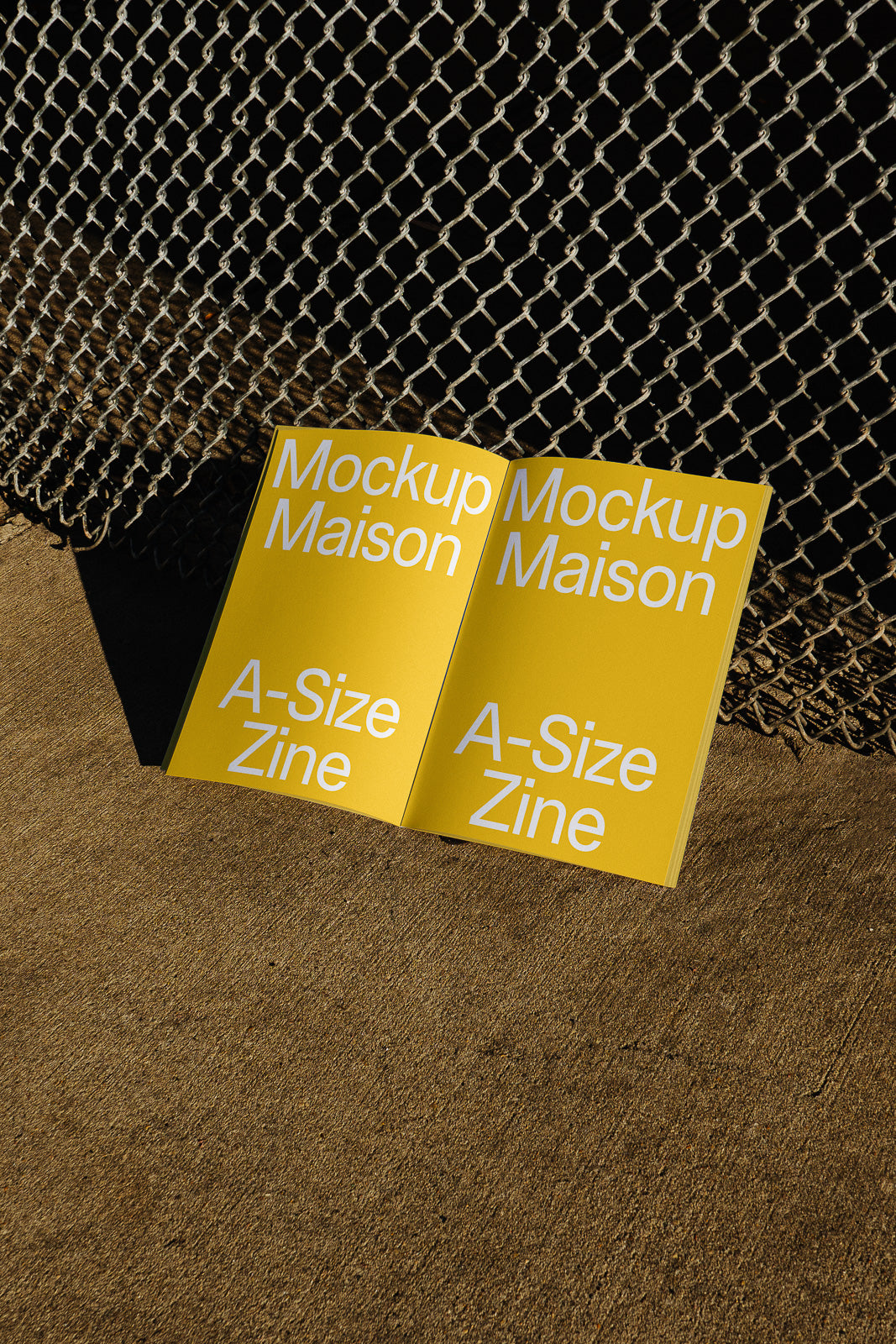 PSD mockup of an A-size zine with yellow pages and bold white text, titled "Mockup Maison," placed on a concrete surface near a chain-link fence.