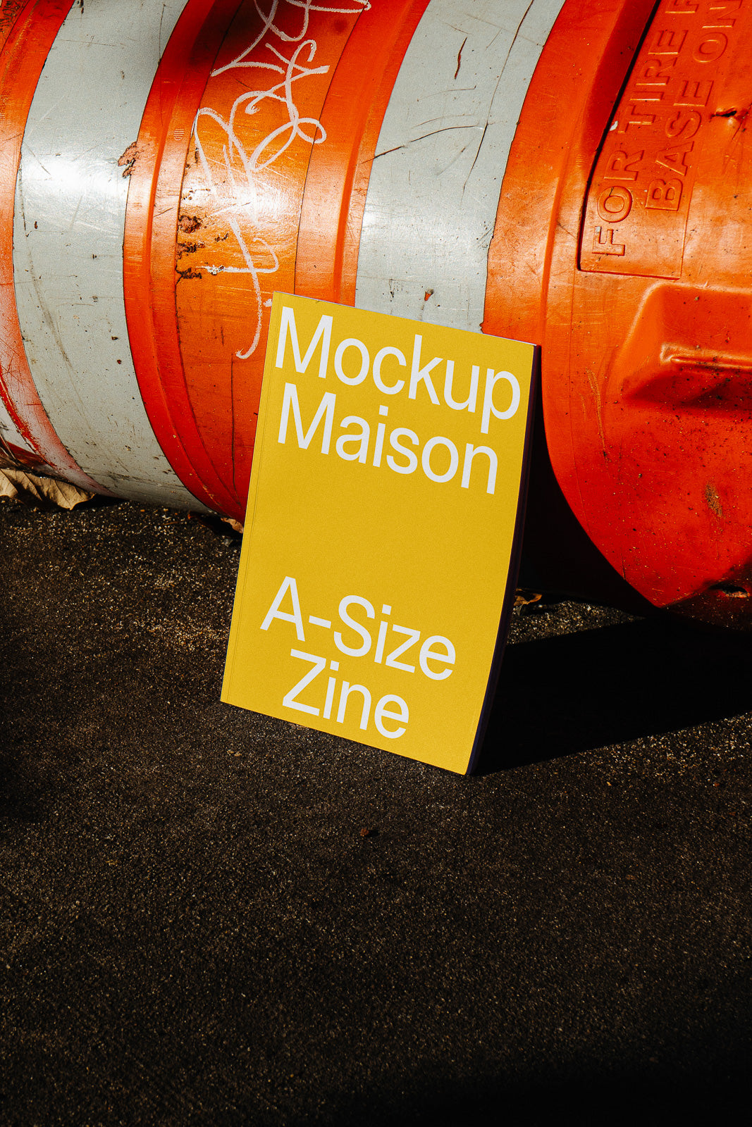 PSD mockup of a yellow A-size zine leaning against an orange and gray construction barrel on a sunny day.