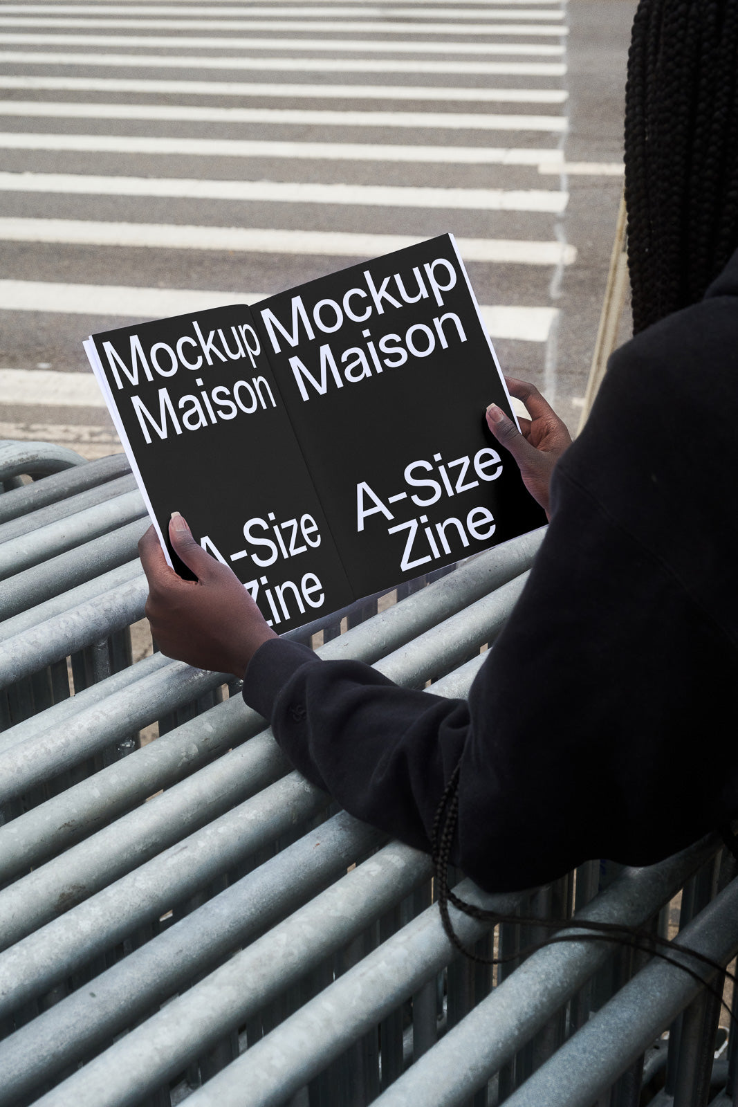 PSD mockup of a person holding an open A-size zine with bold text "Mockup Maison" on both pages, standing by a metal fence near a pedestrian crossing.