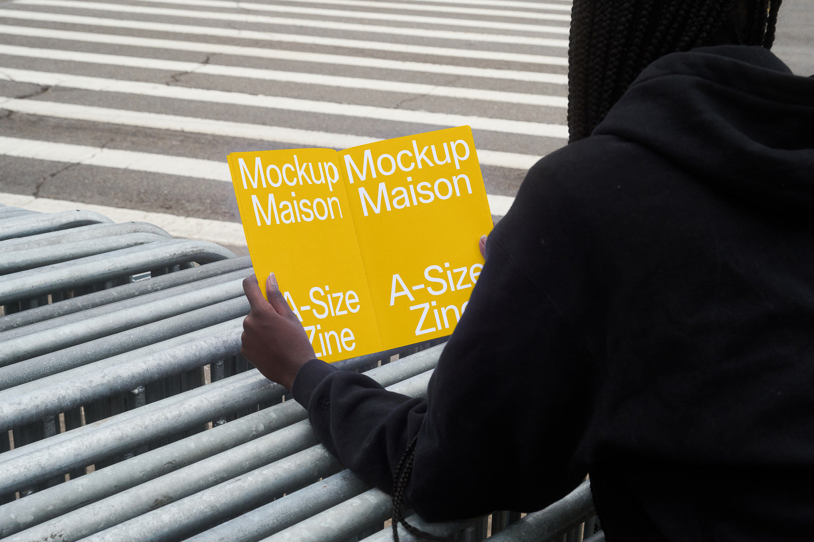 PSD mockup of an open A-size zine held by a person in black clothing, featuring yellow pages with white text, viewed by a crosswalk.