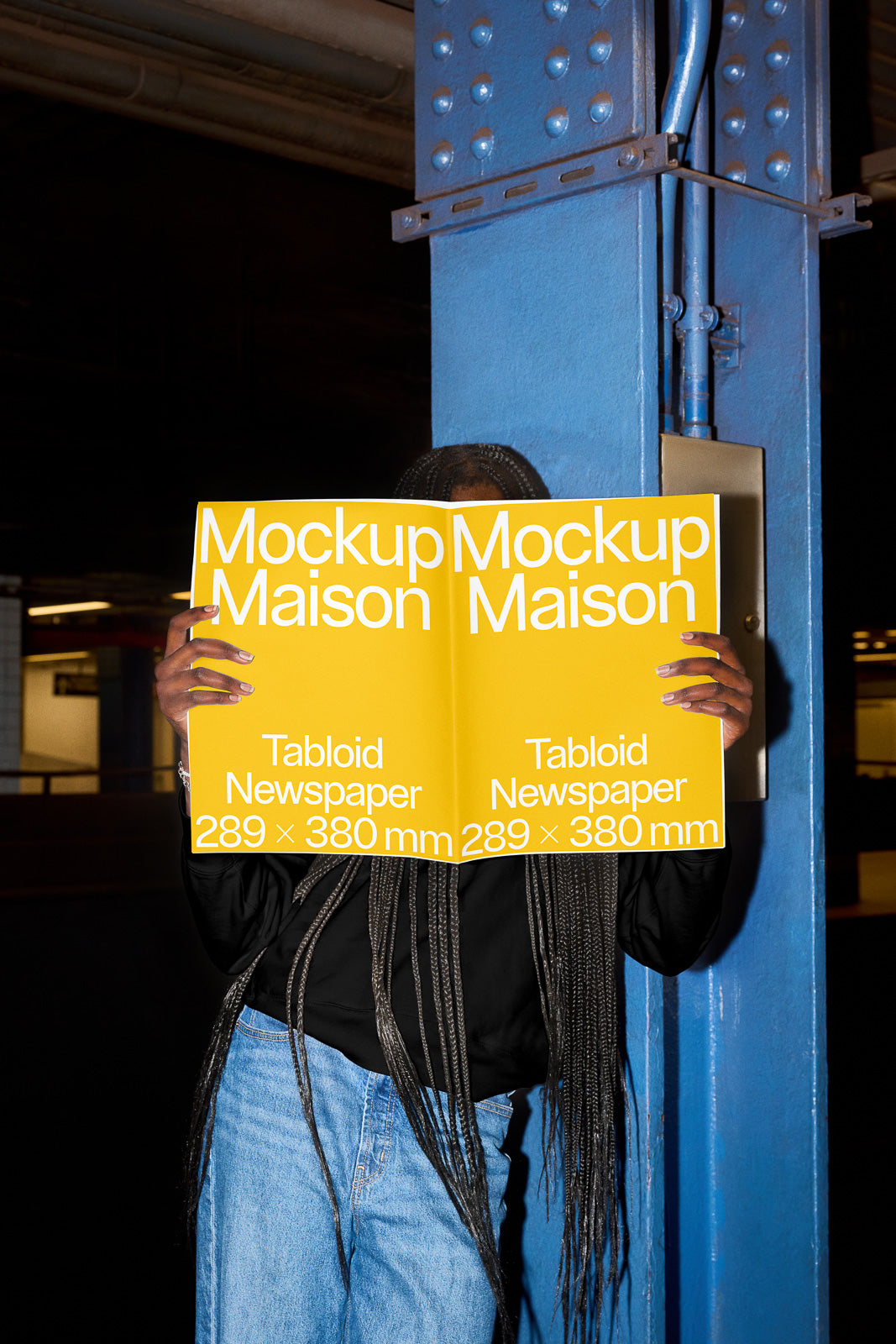 PSD mockup of a person holding a tabloid newspaper, reading "Mockup Maison" with dimensions 289 x 380 mm, against an industrial background.