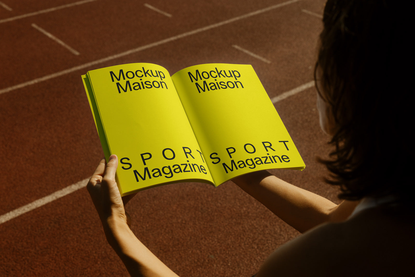 PSD mockup of an open yellow sport magazine held by a person, viewed from above on a running track background.