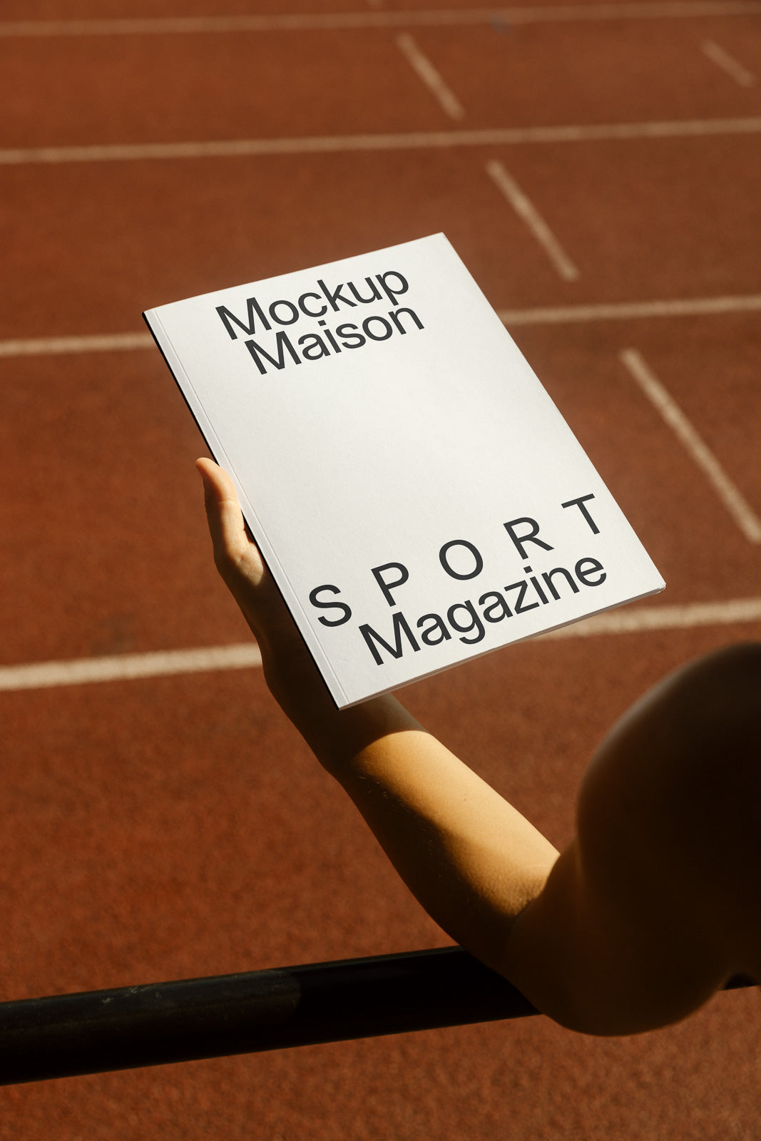PSD mockup of a person holding a magazine titled "Mockup Maison SPORT Magazine" against a background of an athletic track.