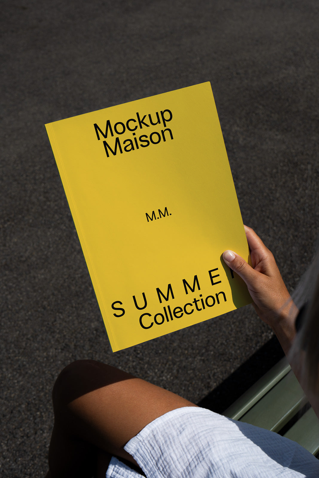 PSD mockup of a person holding a yellow booklet titled "Mockup Maison Summer Collection" with the initials "M.M." visible under bright lighting.