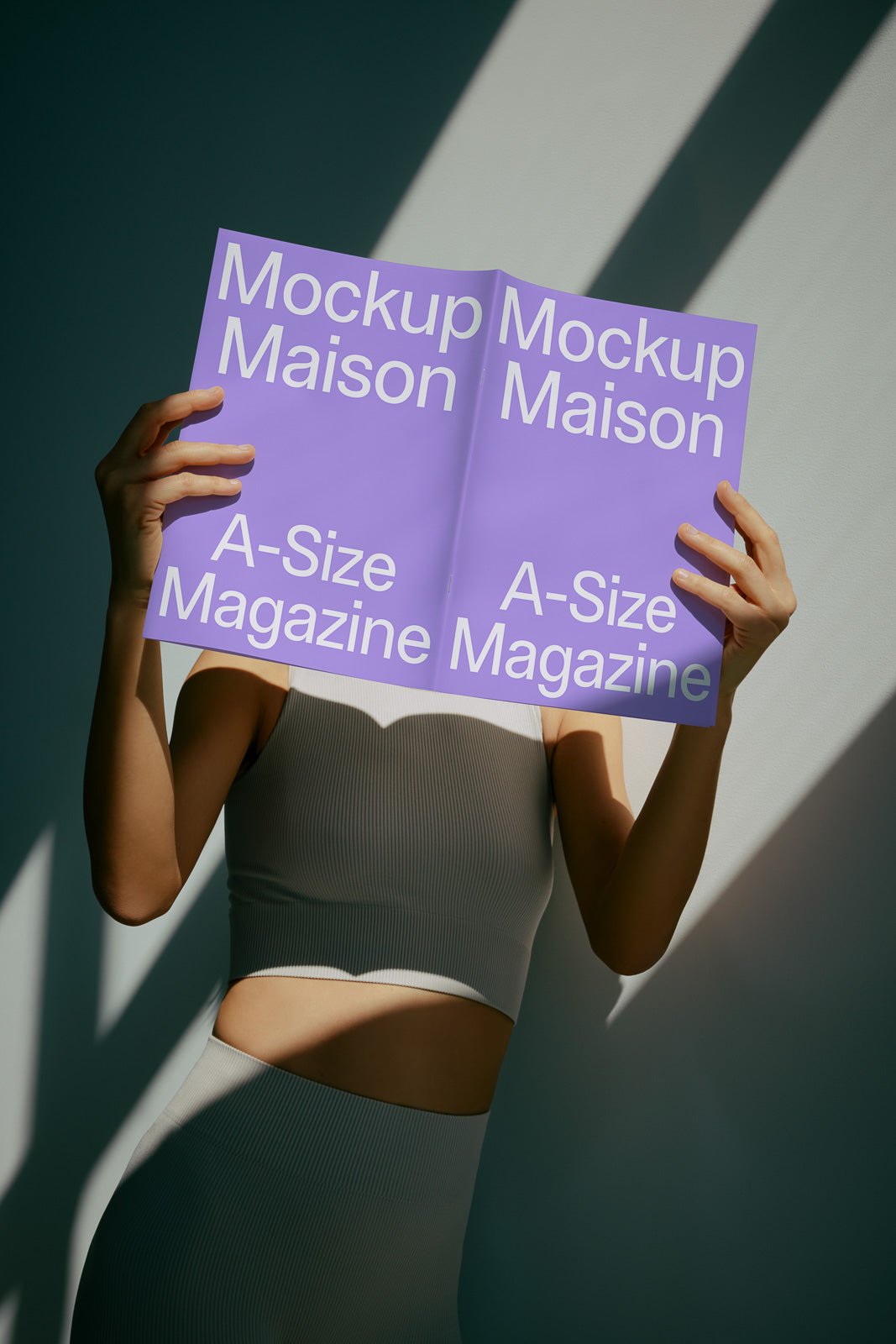 PSD mockup of a person holding an A-size magazine with "Mockup Maison" text, wearing a sleeveless top in dramatic lighting.
