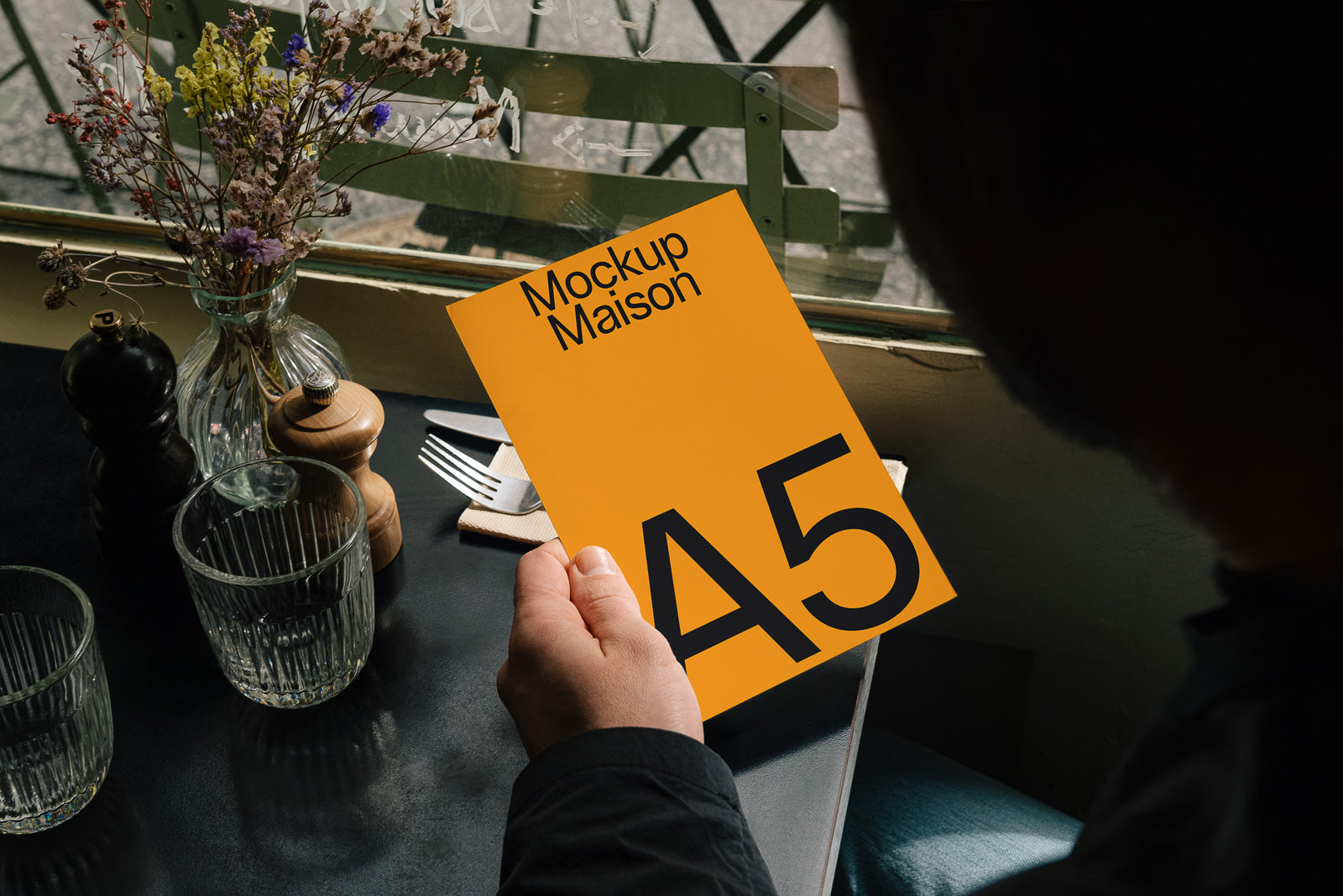 PSD mockup of a person holding an orange A5 card with "Mockup Maison" text, seated at a table with glasses, cutlery, and a flower vase near a window.