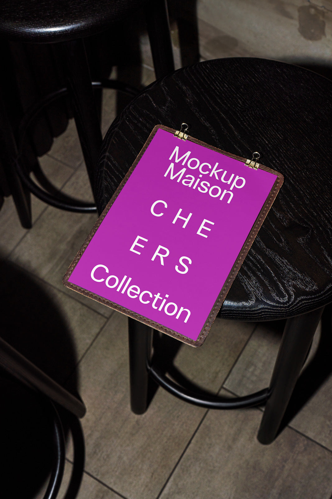 PSD mockup of a clipboard displaying "Mockup Maison CHEERS Collection" text in white on a vibrant pink background, resting on a dark wooden stool.