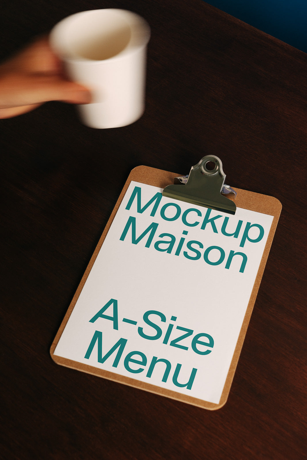 PSD mockup of a clipboard displaying an A-size menu with "Mockup Maison" text, placed on a dark wooden surface. A hand holding a white cup is visible.
