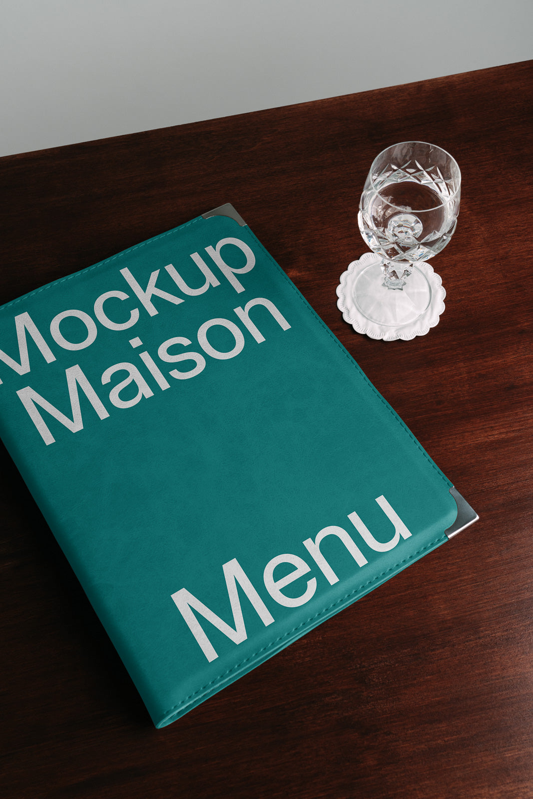 PSD mockup of a teal menu cover on a wooden table with a crystal glass on a coaster beside it.