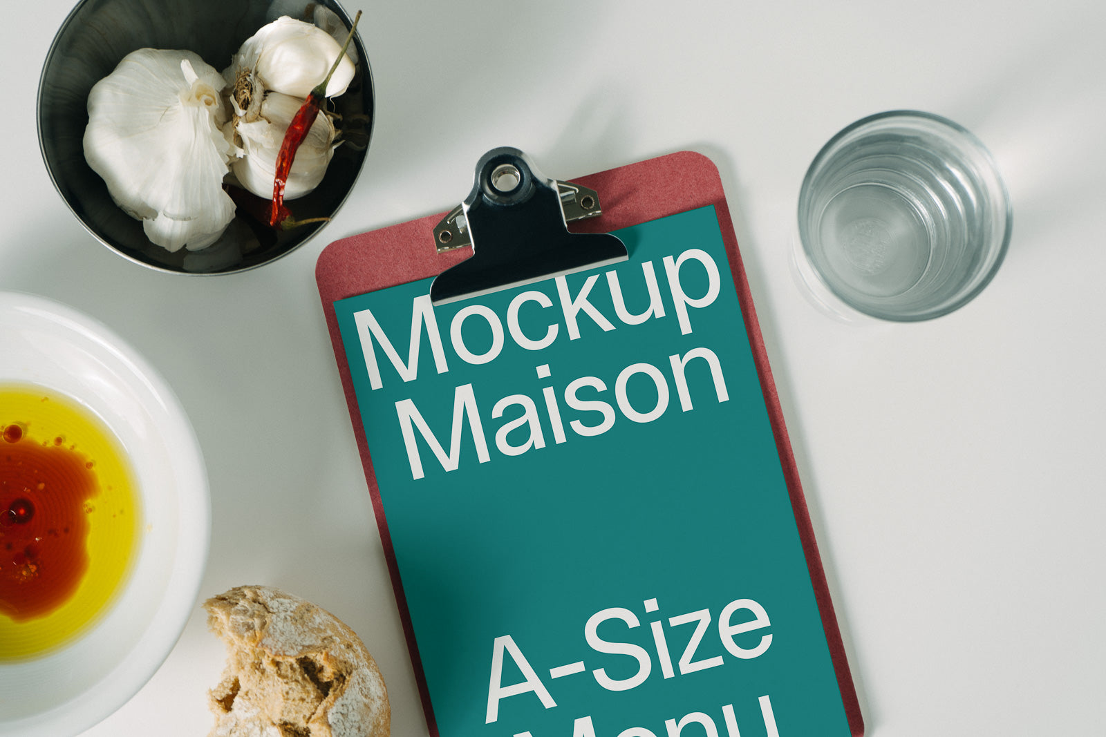 PSD mockup of a red clipboard with "Mockup Maison A-Size Menu" text, surrounded by a bowl of garlic and chili, a glass of water, olive oil, and bread.