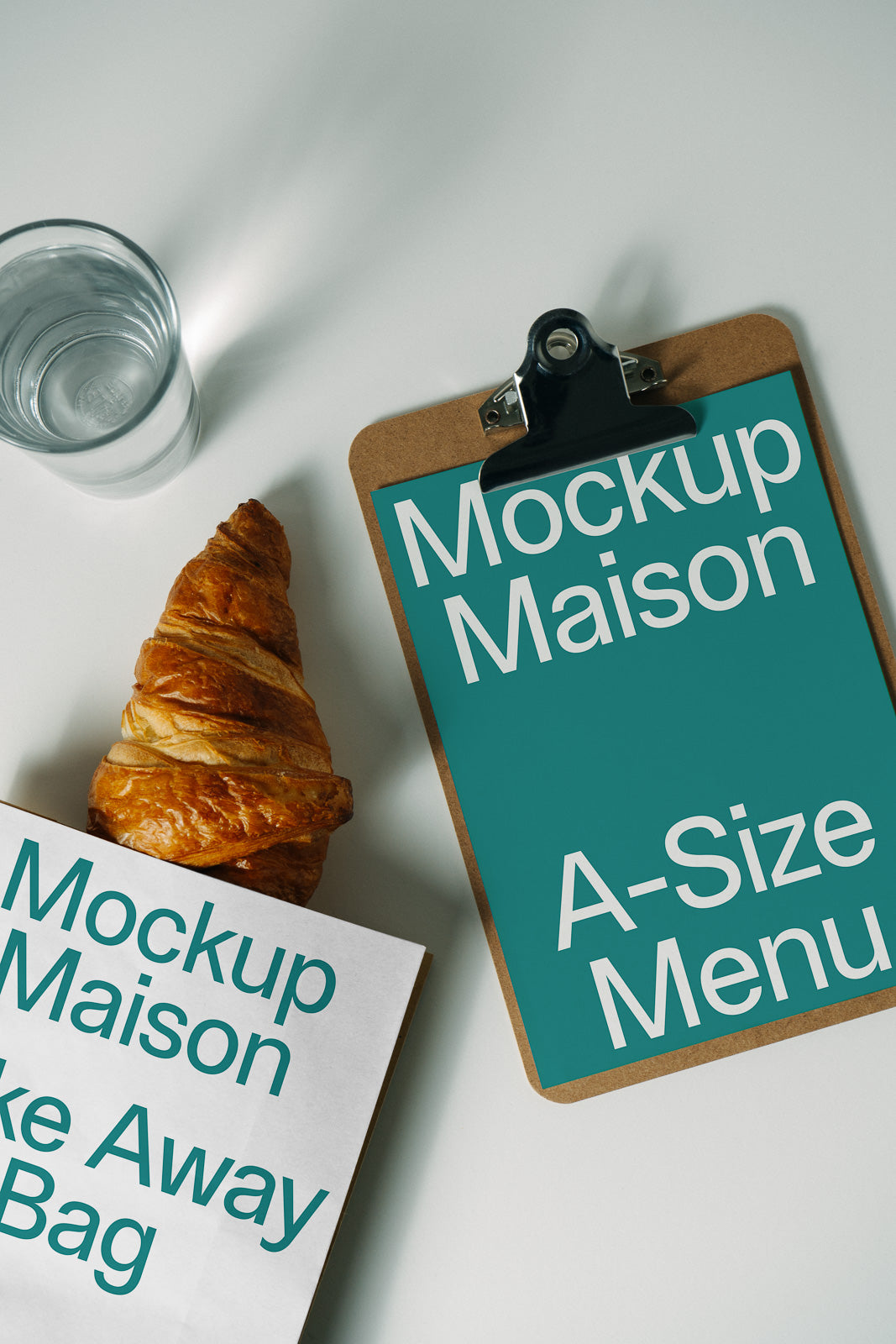 PSD mockup of a green clipboard with "Mockup Maison A-Size Menu" text, next to a takeaway bag labeled "Mockup Maison" and a croissant, with a glass of water.
