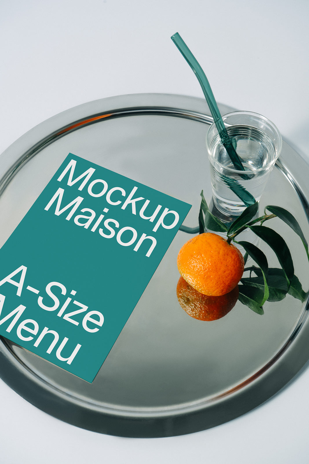 PSD mockup of an A-size teal menu on a round silver tray with a glass of water and a ripe orange, set against a light background.