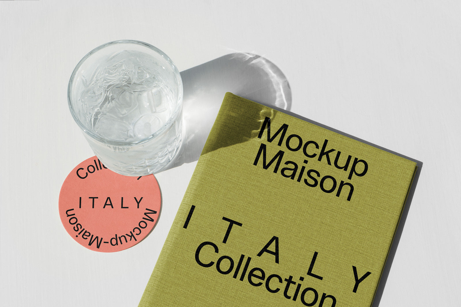 PSD mockup of a glass on a coral-colored coaster beside a green book titled "Mockup Maison Italy Collection" on a white surface.