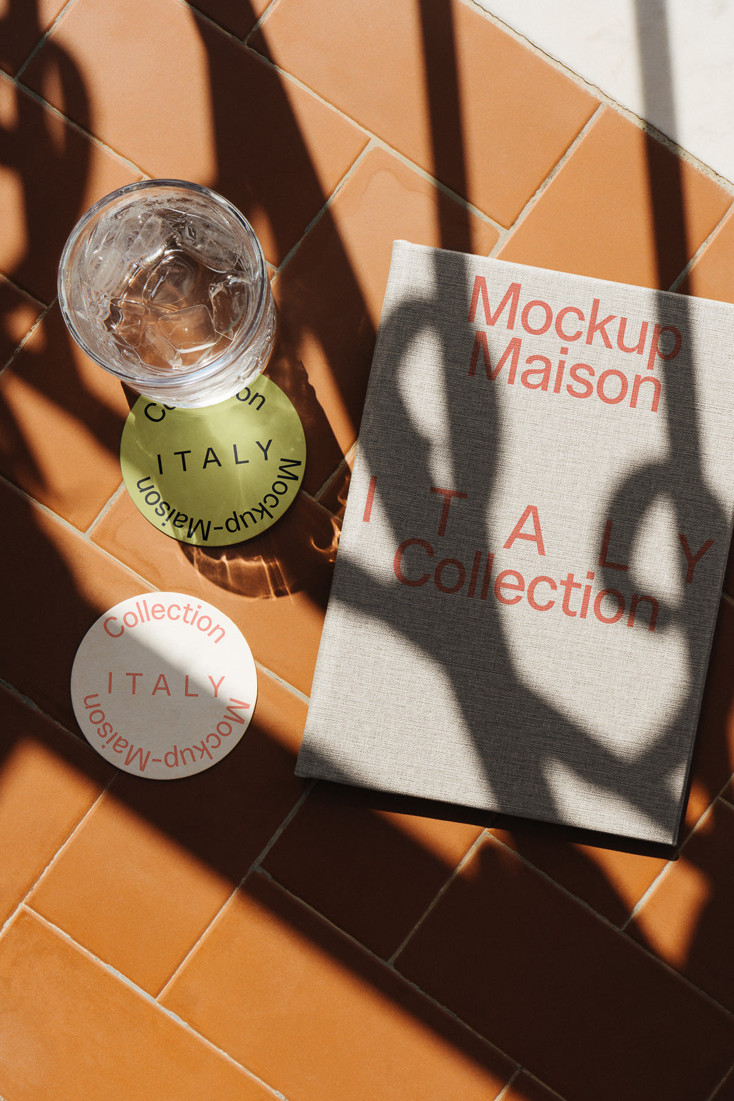 PSD mockup of a hardcover book titled "Italy Collection" alongside coasters and a glass of water on a terracotta tile surface with shadows.