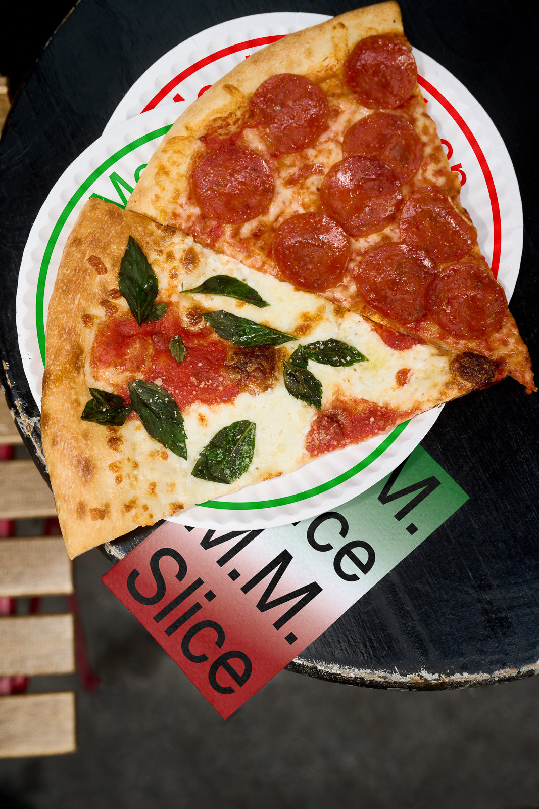 PSD mockup of two pizza slices: one with pepperoni and the other with basil and tomato, placed on a branded paper plate over a wooden surface.