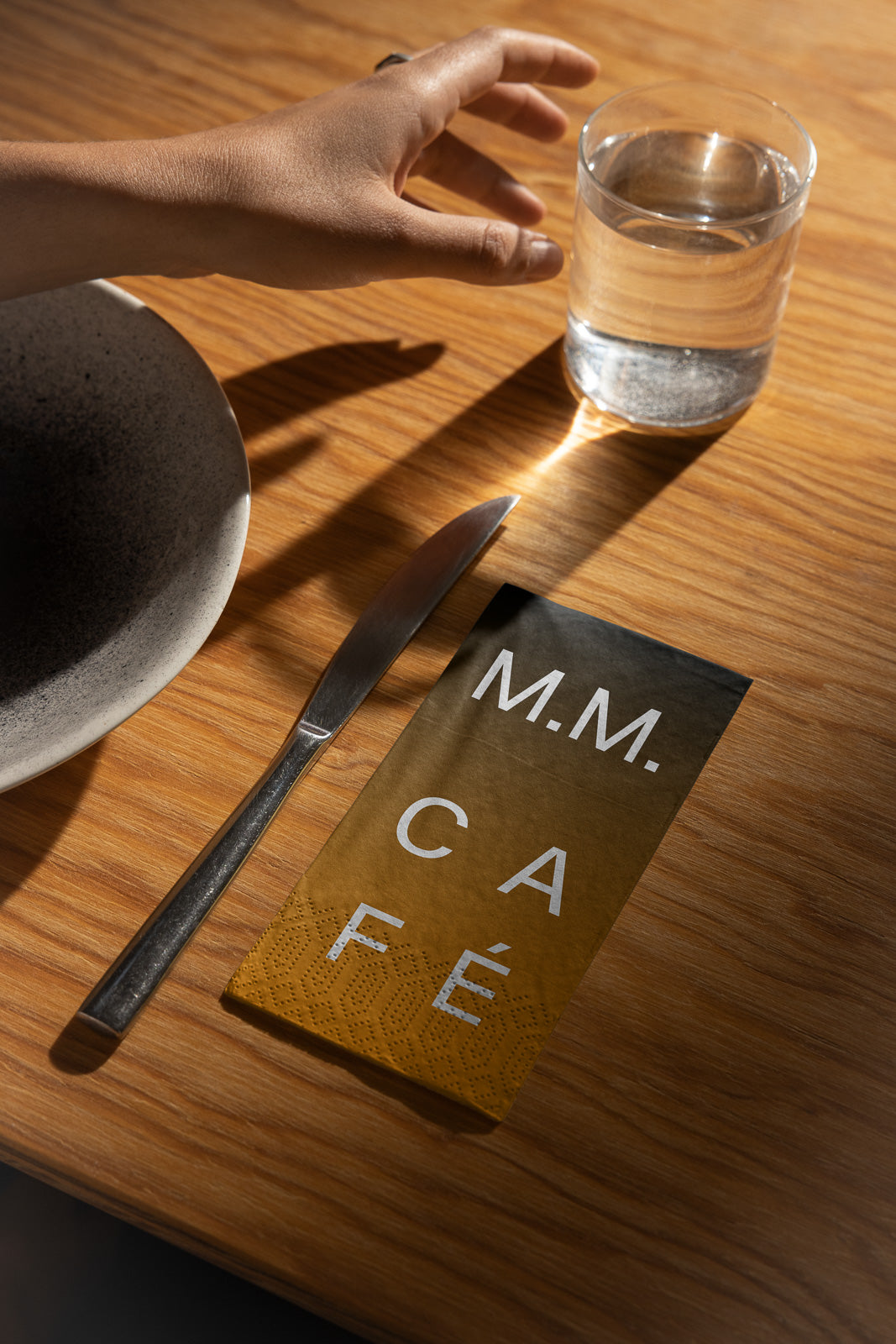 PSD mockup of a café menu on a wooden table with a knife, glass of water, and a hand reaching for the glass, creating a casual dining setting.