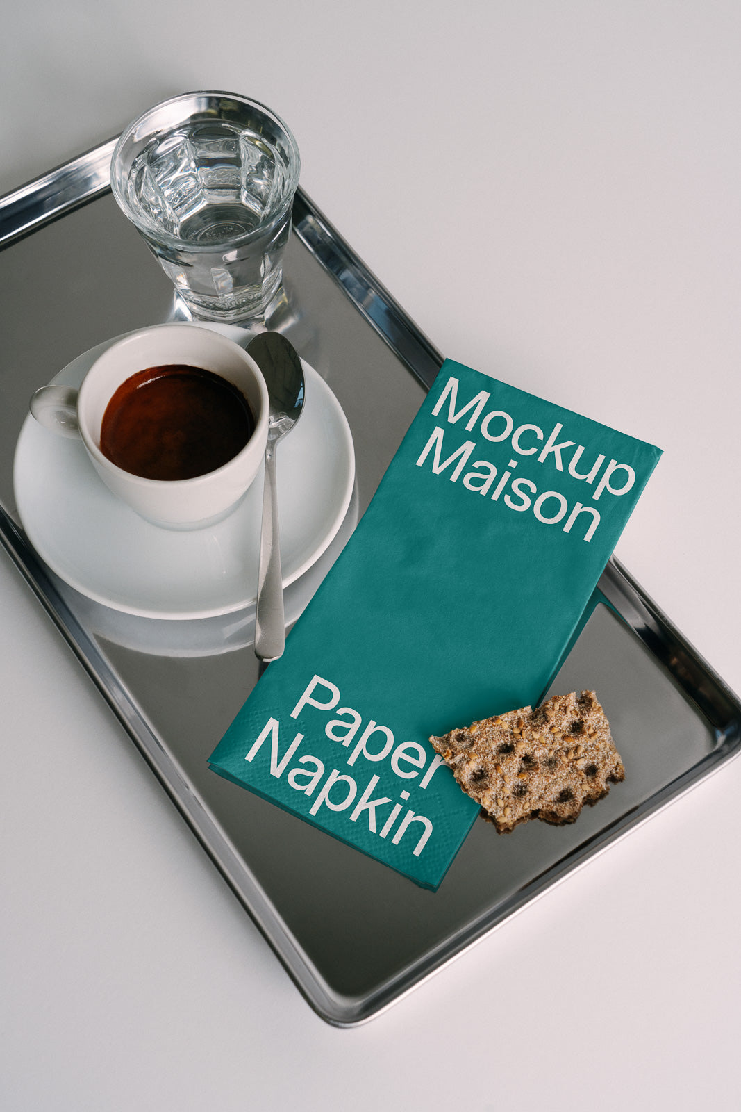 PSD mockup of a teal paper napkin labeled "Mockup Maison" on a metal tray with a cup of espresso, a glass of water, and a piece of crispbread beside it.