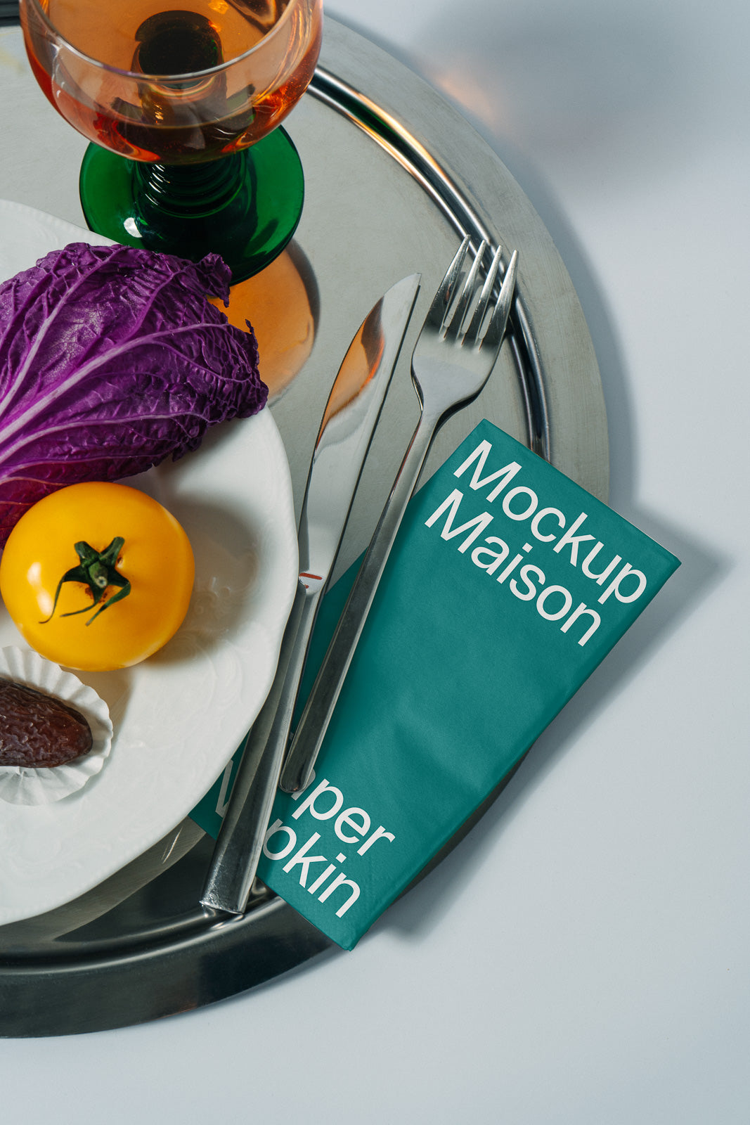PSD mockup of dining setup featuring a white plate with purple cabbage, yellow tomato, and date, framed by silver cutlery and a teal napkin on a tray.