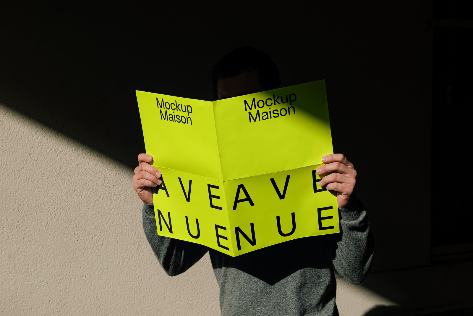 PSD mockup of a person holding a bright green unfolded brochure in a shadowed space, with "Mockup Maison" text at the top and large, bold letters below.