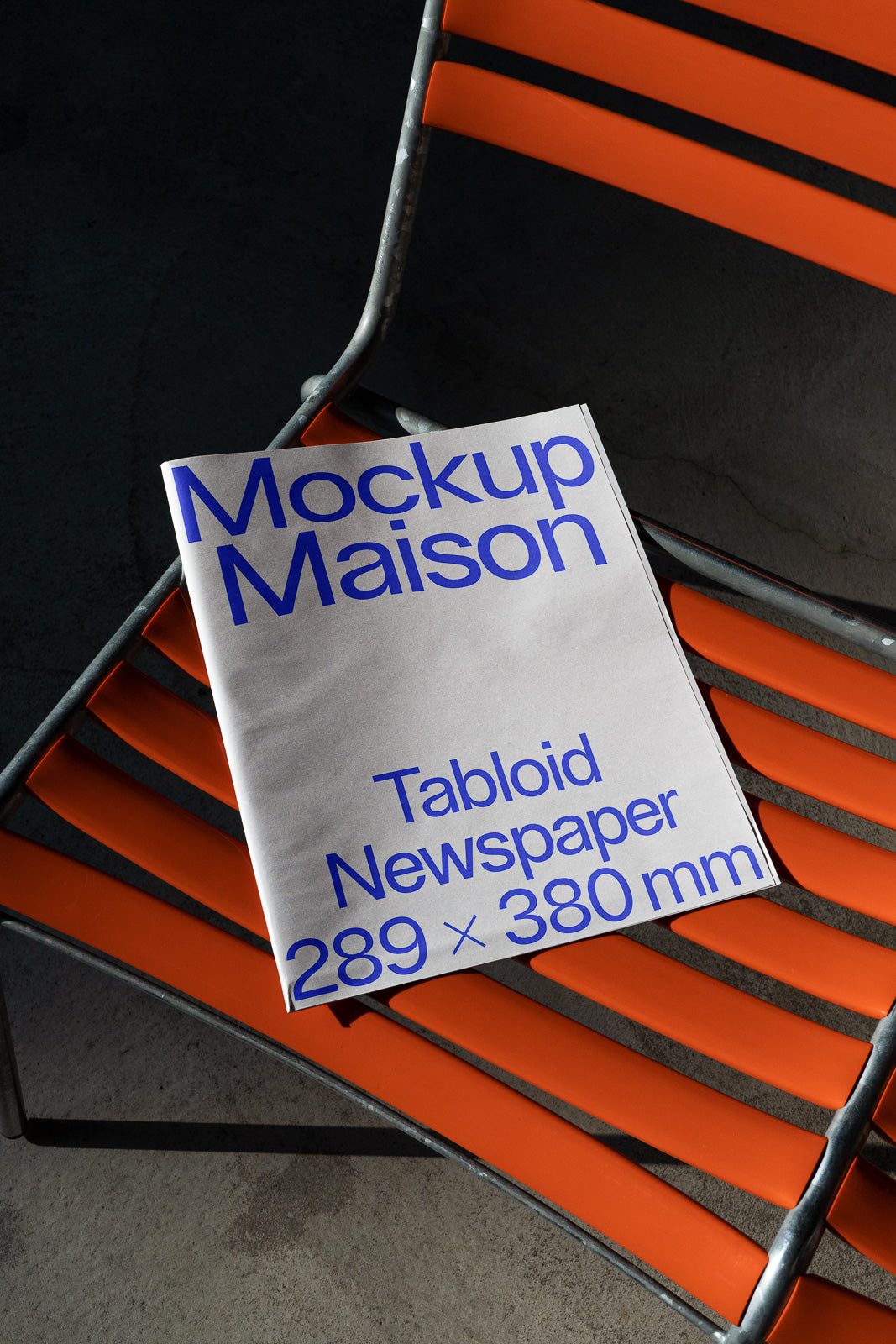 PSD mockup of a tabloid newspaper placed on an orange metal chair, displaying "Mockup Maison" and its dimensions, 289 x 380 mm, in blue text.