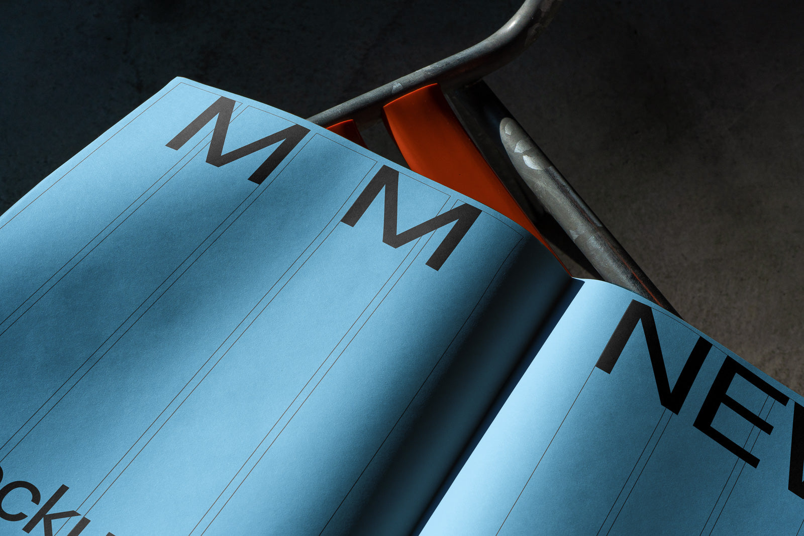 PSD mockup of a blue magazine spread on an orange metal chair, showing large black letters "M" and "NEW" against the page.