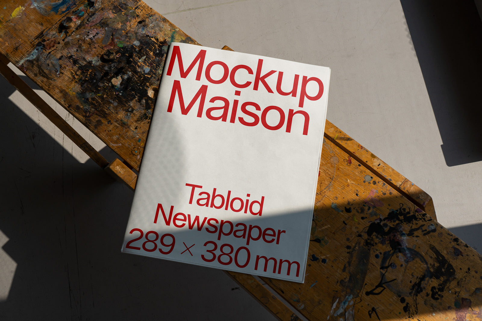 PSD mockup of a tabloid newspaper with red text on a white cover, measuring 289 x 380 mm, resting on a worn, paint-splattered wooden surface.
