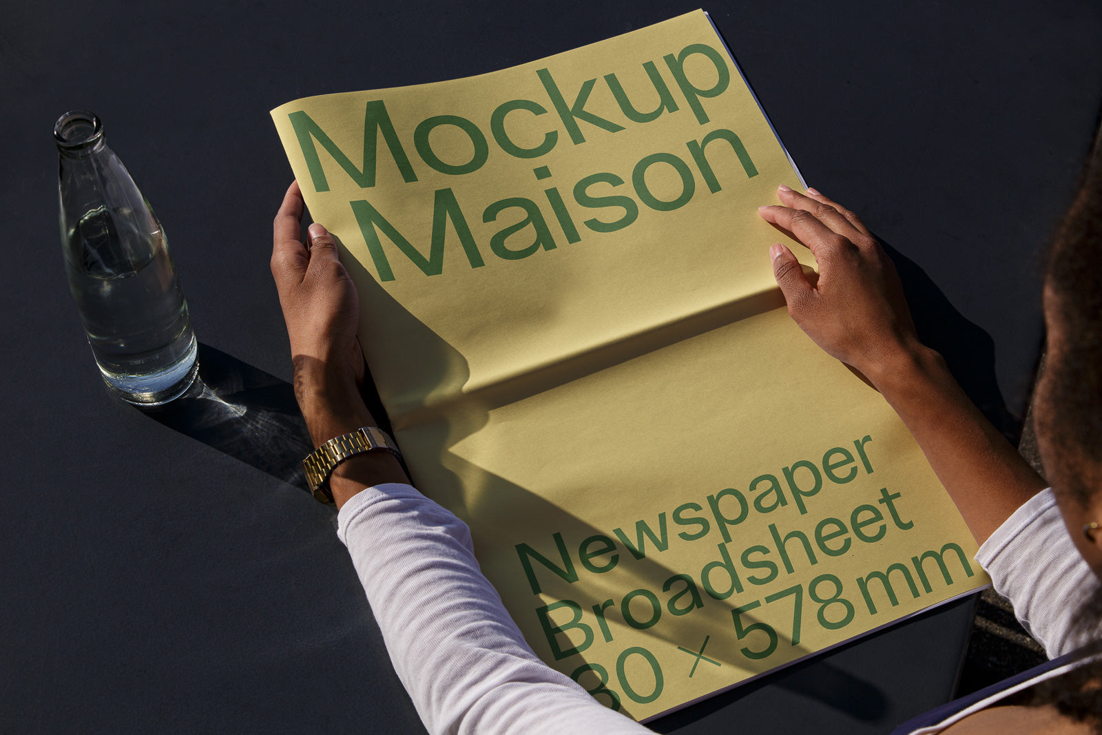 PSD mockup of a yellow broadsheet newspaper titled "Mockup Maison" held open by hands, with a bottle of water on a dark surface.