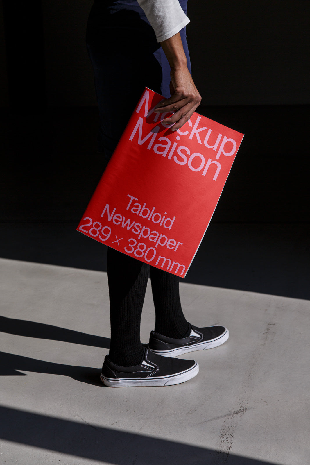 PSD mockup of a person holding a tabloid newspaper with a red cover labeled "Mockup Maison, Tabloid Newspaper, 289 x 380 mm" against a dark background.