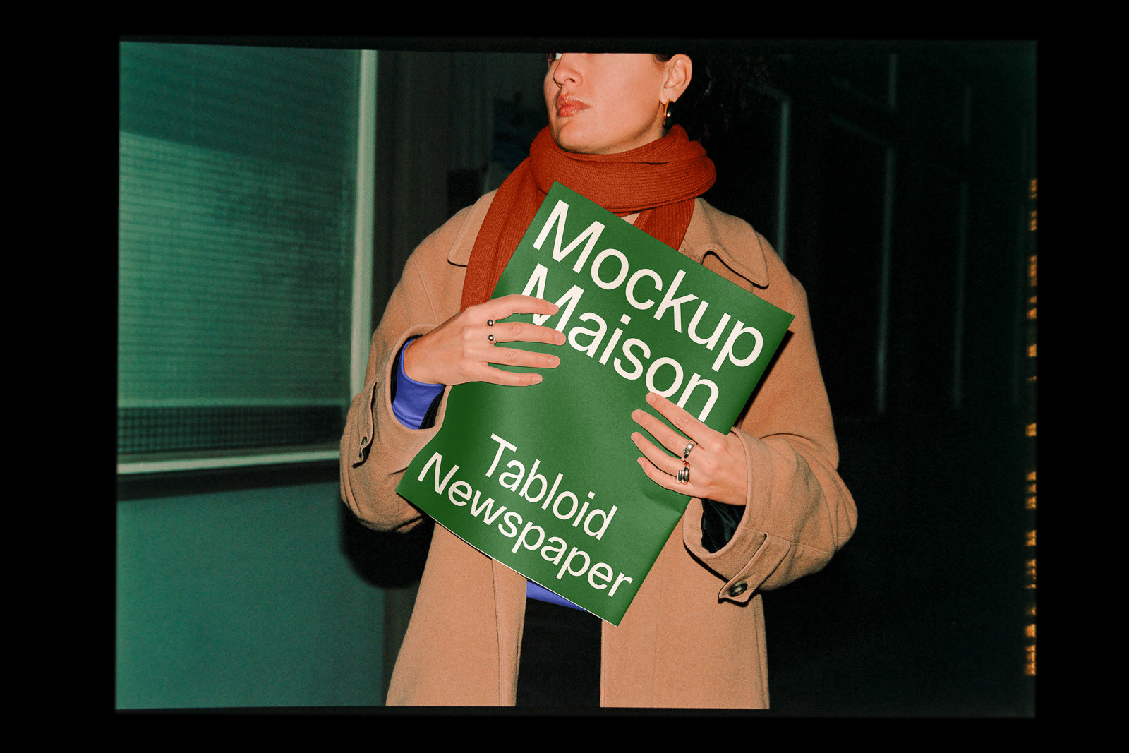 PSD mockup of a person holding a green tabloid newspaper with the words "Mockup Maison" and "Tabloid Newspaper" in white text. The person wears a beige coat.
